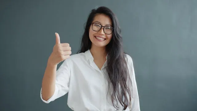 Smiling woman in glasses and white shirt giving a thumbs up against gray background.